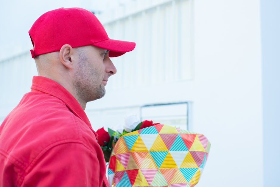 A delivery person dressed in a red cap and matching red jacket holding a large floral gift wrapped in colourful geometric patterned paper with shades of yellow, blue, red, and orange. The flowers insi