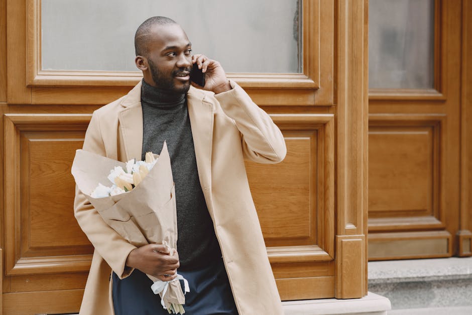 A smiling man with a beard, dressed in a beige coat and black turtleneck, is standing outdoors in front of a wooden-paneled building. He is holding a bouquet of fresh white and cream flowers wrapped i