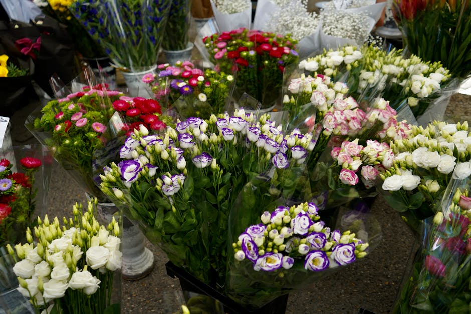 An array of fresh floral arrangements displayed on a dark surface inside a flower shop, featuring bunches of carnations in shades of white, pink, purple, and red, all wrapped in clear cellophane. The