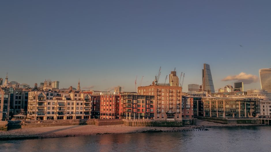 A scenic view of the Thames River with a lineup of modern and historic buildings along the Blackfriars area in London during golden hour, featuring a clear sky with soft sunlight casting warm tones on