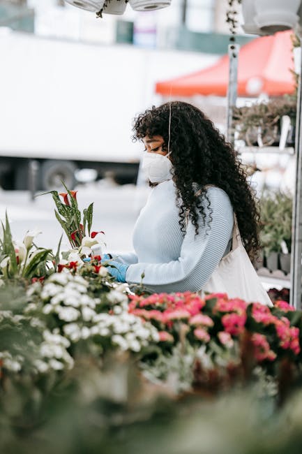 A woman with curly dark hair, wearing a white face mask, light blue sweater, and gloves, is arranging a bouquet of fresh flowers on an outdoor market stand. The bouquet includes red roses, white lilie